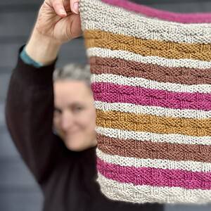 A white woman with brown eyes and silver hair is standing in front of a painted wooden wall and looking at the camera. She wears a black sweater. She is in the background of the photo and is out of focus. She is holding up a knitted cowl to the foreground in stripes of pale grey, ochre, brown and fucshia. The cowl has a basketweave texture.