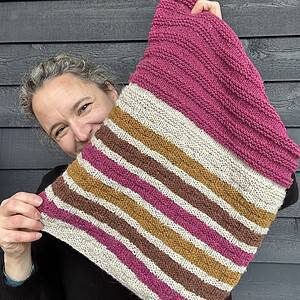 A white woman with brown eyes and silver hair is standing in front of a painted wooden wall and looking at the camera. She is smiling widely though her mouth is hidden behind the cowl she is holding in front of her. She wears a black sweater. She is holding up a knitted cowl which has basketweave textured stripes of pale grey, ochre, brown and fucshia at the bottom and solid fuchsia pink with garter stitch stripes for texture at the top.